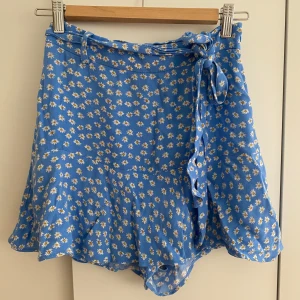 Blommig blå omlottkjol från Zara - Supersöt blå kortkjol från Zara med vita och gula prästkragar, omlottmodell med knyt i midjan och volangig nederkant. Kjolen inbyggda shorts som kan ses på sista bilden.