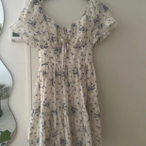 Blommig kortklänning med puffärm - Supersöt kortklänning i ljusbeige med blått blommönster. Klänningen har puffiga korta ärmar, knytning framtill och volang i kjolen. Perfekt för sommarens alla dagar och ger en romantisk vibe.