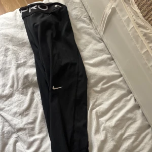 Nike Pro svarta tights  - Svarta Nike Pro tights med hög midja och bred resår med logga. Tight passform och stretchigt material som passar perfekt till träning. Klassisk Nike Swoosh på benet. Snygga och bekväma för dig som gillar sportig stil.