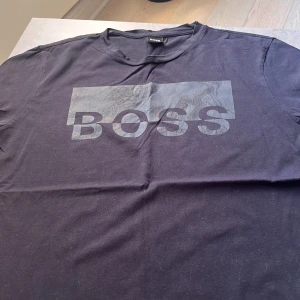Svart BOSS t-shirt med stor logga - Svart t-shirt från BOSS med stor, diskret logga i grått över bröstet. Klassisk rund halsringning och korta ärmar. Tillverkad i mjuk bomull för en skön känsla och enkel stil. Storlek Large.
