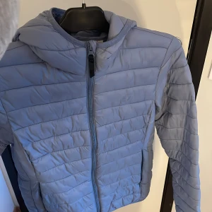 Ljusblå pufferjacka från Hangten - Säljer en ljusblå pufferjacka från Hangten i storlek S. Jackan har huva, dragkedja framtill och två sidofickor. Den quiltade designen ger en clean och modern look, perfekt för kyliga dagar. Enkel att matcha med olika outfits.