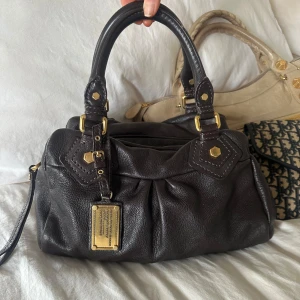 Mörkbrun Q Classic Baby Groove väska! - Så snygg mörkbrun klassisk handväska i nyskick från Marc by Marc Jacobs i äkta läder med gulddetaljer och dubbla handtag. Absolut snyggaste färgen som passar till allt, den perfekta sommarväska än som rymmer mycket men är smidig och nätt. Lilla modellen Q Classic. Inga repor eller hål på väskans läder. Otroligt välbevarad vintage modell! Perfekt för att ge en stilren touch till din outfit. Skriv privat för fler bilder. Mått 34x18x12 (undersida). Kan hämtas hemma hos mig i Nacka eller skickas🤍