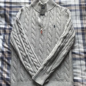 Grå kabelstickad tröja Polo Ralph Lauren - Kabelstickad grå tröja från Polo Ralph Lauren med halv dragkedja och klassisk broderad logga på bröstet. Tröjan har hög krage, ribbade muddar och är långärmad. Perfekt för lager-på-lager och snygg till jeans.