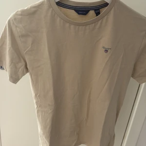 Beige t-shirt från GANT - Snygg beige t-shirt från GANT med liten broderad logga på bröstet. Klassisk rund halsringning och korta ärmar. Tillverkad i mjuk bomull som känns skön mot huden. Perfekt för en clean och stilren look. Strl är i vuxen storlek XS. Skick 8,5/10