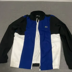 Lacoste Sport vindjacka blå/svart/vit - Snygg vindjacka från Lacoste Sport ÄKTA! I blått, svart och vitt med dragkedja framtill. Jackan har lång ärm, klassisk krage och coolt Lacoste-tryck på ryggen. Tillverkad i polyester, perfekt för dig som gillar sportig och stilren look.