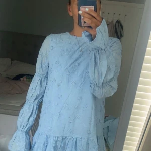 Blå broderad klänning med öppen rygg - Supersöt ljusblå klänning med broderade blommor, volang nertill och långa puffärmar. Öppen rygg med knytband upptill ger en extra snygg detalj. Perfekt för sommarens alla tillfällen! Använd vid ett tillfälle