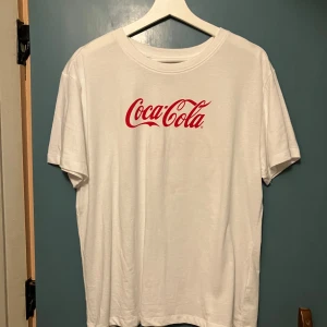 Vit Coca-Cola t-shirt  - Vit t-shirt  med klassiskt Coca-Cola-tryck i rött framtill och stort retrotryck med flaska och texten 'Always Coca-Cola' på ryggen. Skön passform, rund hals och korta ärmar. Perfekt för dig som gillar ikoniska loggor och streetstyle.