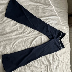 Mörkblå bootcut träningsbyxor Muséra - Mörkblå bootcut träningsbyxor från Muséra Sport med vita detaljer längs midjan. Tillverkade i stretchigt material med 75% polyester och 25% elastan, vilket gör dem perfekta för träning. Snygg passform med hög midja och diskret logga framtill.