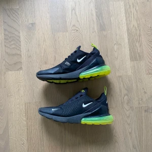 Nike Air Max 270 Svarta Sneakers - Svarta Nike Air Max 270 sneakers med vit logga och neongul detalj på sulan. Skorna har en luftig mesh-överdel, snörning och en stor synlig Air-enhet i hälen för extra komfort. Perfekta för dig som gillar sportig och modern stil. Dem saknar däremot snören.