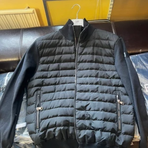 Svart quiltad pufferjacka Moncler - Snygg svart pufferjacka från Moncler med quiltad framsida och släta ärmar. Jackan har dragkedja framtill, två fickor med dragkedja och ribbade muddar. Materialmix av polyester och bomull ger både värme och stil. Perfekt för dig som vill ha en clean och modern look.