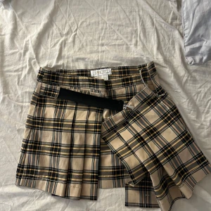 Rutig kortkjol från Zara TRF - Superfin rutig kortkjol från Zara TRF i beige, svart och gula toner. Kjolen har klassiska veck, omlott-detalj och ett svart bälte med spänne framtill. Perfekt för en trendig och preppy look. Materialet är polyester och rayon.