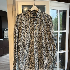 Blommig skjorta från Sand Copenhagen - Snygg skjorta från Sand Copenhagen med blommigt mönster i blått och vitt. Skjortan har lång ärm, klassisk krage och knäppning framtill. Modern fit som sitter stilrent och passar perfekt till dig som vill sticka ut lite extra nu på midsommar. Inköpt på Carl XI Mode i Karlskrona 2021 och endast använd två gånger. Nypris 1400kr