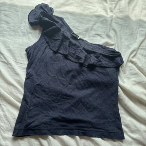 Mörkblå oneshoulder topp Add It - Snygg mörkblå oneshoulder topp från Add It med volangdetalj längs halsringningen. Toppen är ärmlös på ena sidan och har en cool asymmetrisk design som sticker ut. Perfekt för dig som vill ha något unikt och trendigt i garderoben.