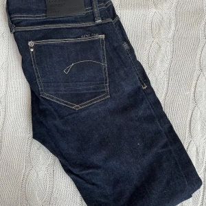 Mörkblå jeans från G-Star RAW GS01 strl 30/32 - Snygga mörkblå jeans från G-Star RAW med klassisk femficksdesign och kontrastsömmar. Jeansen har raka ben och tydliga logodetaljer på bakfickan och myntfickan. Perfekta för dig som gillar stilrena och tidlösa denimplagg.
