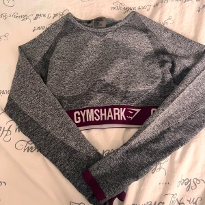 Grå croppad tröja från Gymshark - Snygg gråmelerad croppad tröja från Gymshark med långa ärmar och bred elastisk lila resår med logga nedtill. Perfekt för träning eller chill, med sportig vibe och bekväm passform.