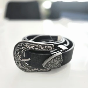Svart westernbälte från Factorie - Snyggt svart bälte från Factorie med bred westerninspirerad silverspänne och detaljerade blommönster i metall. Bältet är gjort i 100% polyuretan och har en cool, stilren look som lyfter vilken outfit som helst. Passar midjemått upp till 92 cm.