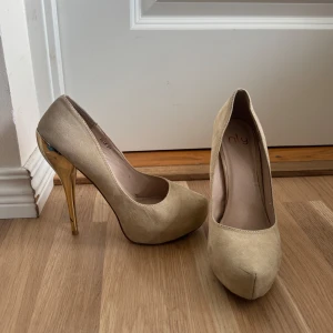 Beige pumps med guldklack från NLY - Stilrena pumps i beige mocka-look från NLY med höga, smala klackar i metallicguld. Skorna har rund tå och plattform framtill för extra höjd. 