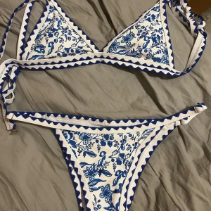 Blåvit blommig bikini  - ALDRIG ANVÄND!! Superfin bikini i blått och vitt med blommigt mönster och coola sicksack-detaljer längs kanterna. Triangeltopp med spänne i ryggen samt matchande trosa med smala band. Perfekt för stranden eller poolen i sommar!