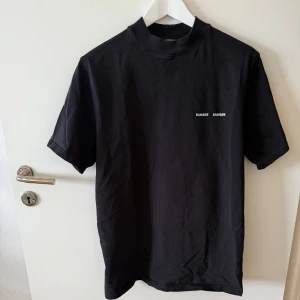 Svart t-shirt från Samsøe Samsøe - Svart t-shirt från Samsøe Samsøe med liten vit logga på bröstet. Klassisk modell med korta ärmar och ribbad högre halsringning. Perfekt basic-plagg i mjuk bomull som funkar till allt.