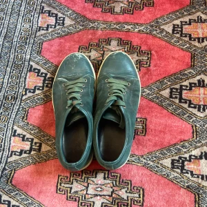 Gröna sneakers från Lanvin i skinn - Lanvin sneakers i mörkgrönt skinn med vita sulor och klassisk snörning. Skorna har rund tå och en stilren, minimalistisk design. Perfekta för dig som gillar exklusiva och tidlösa sneakers med en unik färg. På toe capen så är de lite slitet men går att fixa tror jag via sko makar med ny färgning, står storlek 8 på den men passar mig som har storlek 40-41