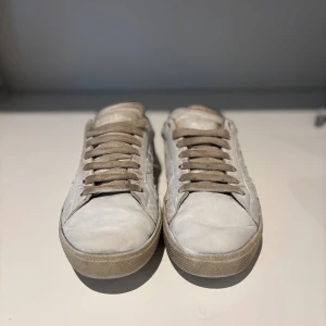 Saint Laurent vita sneakers med stjärnor - Snygga vita sneakers från Saint Laurent med grå snörning och coola stjärndetaljer på sidorna. Om du gör rent skosnaörna så blir de som nya. Skorna har en beige sula och svartgrå hälparti med logga. Tillverkade i skinn och har en klassisk rund tå och platt sula. Litet hål vid en sjärna skriv om ni vill ha bild spelar ingen roll egentligen inget vatten kommer in eller något