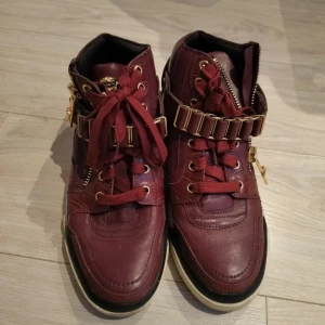Vinröda Versace sneakers med gulddetaljer - Exklusiva vinröda sneakers från Versace i skinn med feta gulddetaljer, dragkedjor och snörning. Snygg Medusa-logga på plösen och Versace-tryck bak på sulan. Skorna har en chunky sula i svart, vitt och guld. Perfekt statement-piece för dig som vill sticka ut.