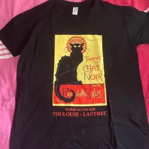 Svart Chat Noir t-shirt med tryck - Svart t-shirt med ikoniskt Chat Noir-tryck i gult, rött och svart framtill. Klassisk passform och rund hals. Perfekt för dig som gillar vintage och fransk konststil. Storlek XS men passar mer som liten M eller stor S