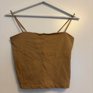 Beige linnetopp från Gina Tricot - Säljer en enkel och stilren brun/beige linnetopp från Gina Tricot. Toppen har tunna axelband och rak skärning, perfekt för varma dagar. Tillverkad i mjuk bomull som känns skön mot huden. Passar dig som gillar minimalistisk och clean stil.