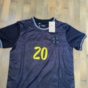 Svart Brasilien Vini Jr. Nike tröja - SELGER FRA NORGE OG ER I NORSKE KRONER!Snygg svart Brasilien fotbollströja från Nike med VINI JR. och nummer 20 i gult på ryggen. Tröjan har korta ärmar, broderat CBF-märke och Nike-logga, samt grå detaljer på krage och ärmslut. Tillverkad i Dri-FIT-material som andas.