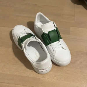 Valentino Garavani Open sneakers vit/grön - Snygga Valentino Garavani Open sneakers i vitt skinn med grön detalj över snörningen. Klassisk rund tå, platt sula och ikoniska nitar baktill. Perfekta för dig som vill sticka ut med exklusiva och trendiga sneakers.