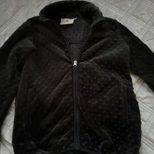 Svart fleecejacka från okänd butik - Mysig svart fleecejacka med coola upphöjda prickar i tyget. Jackan har dragkedja framtill, hög krage och två fickor. Perfekt för dig som gillar en mjuk och chill stil under kyliga dagar.