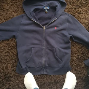 Mörkblå hoodie från Polo Ralph Lauren - Snygg mörkblå hoodie från Polo Ralph Lauren med dragkedja och huva. Klassisk röd logga broderad på bröstet och stora fickor framtill. Perfekt för chill dagar eller när du vill ha en enkel men stilren look.