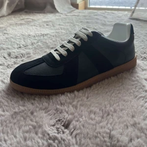 Maison Margiela svarta sneakers i skinn - Maison Margiela sneakers i svart skinn och mocka med vita snören och klassisk gummisula. Snygg retrostil med rund tå och lågt skaft. Perfekta för dig som gillar minimalistisk och stilren design. skriv vid minsta intresse 🙋🏽‍♂️💯🫱🏻‍🫲🏽pris kan diskuteras vis snabb affär 🫱🏻‍🫲🏽💯