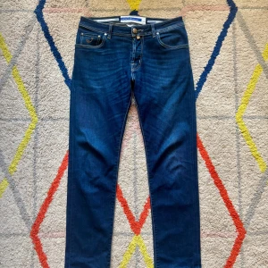 Blå jeans från Jacob Cohen - Snygga blå jeans från Jacob Cohen med klassisk femficksdesign och kontrastsömmar. Byxorna har en normal passform och stängs med knapp och dragkedja. Märkespatch baktill i midjan och diskret broderad J på fickan.