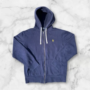 Marinblå hoodie från Polo Ralph Lauren - Marinblå hoodie från Polo Ralph Lauren med dragkedja, vit snörning i huvan och klassisk gul broderad logga på bröstet. Tröjan har långa ärmar, känguruficka och är gjord i mjuk bomull. Perfekt för en chill och stilren look.