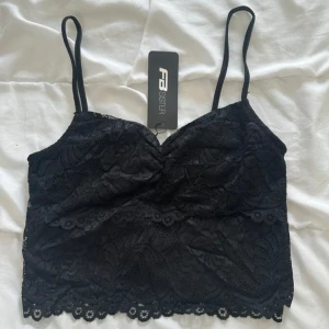 Svart spetsig linnetopp från FB Sister - Supersnygg svart linnetopp från FB Sister med tunna justerbara axelband och vacker blommig spets över hela toppen. Croppad modell med vågig spetskant nertill och lätt vadderad byst. Perfekt att styla med jeans eller kjol för en trendig look