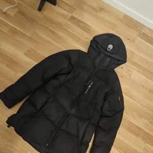 Svart dunjacka Sail Racing med huva - Svart dunjacka från Sail Racing med stor huva och logga framtill. Jackan har dragkedja hela vägen, quiltad design och justerbar nederkant. Perfekt för kalla dagar och har en clean, sportig look.