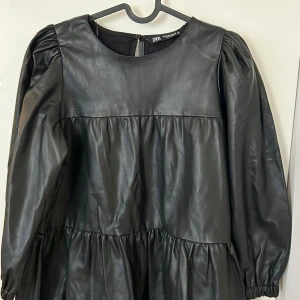 Svart klänning i skinnimitation från Zara - Cool svart klänning från Zara i skinnimitation med puffiga långärmar och rund halsringning. Klänningen har en avslappnad passform och volangdetaljer som ger en trendig look. Perfekt för dig som vill sticka ut med en edgy stil.