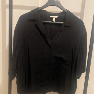 Svart skjorta från H&M i storlek L - Svart skjorta från H&M i storlek L. Skjortan har klassisk krage, knappar framtill och en bröstficka. Modellen är lite loose och har lång ärm. Perfekt att styla med jeans eller dressade byxor.
