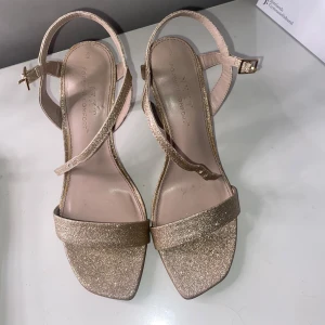Glittriga guldiga klackskor - Snygga sandaler från Next i glittrigt guld med smala remmar och fyrkantig tå. Skorna har hög klack och justerbart spänne i guld. Storlek 38, endast använd 1 gång