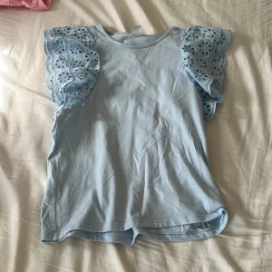 Ljusblå blus med broderade volangärmar - Supersöt ljusblå blus med broderade volangärmar i bomull. Ärmarna har fina hålbroderier ger en trendig vibe. Perfekt för dig som vill ha något extra till din outfit!