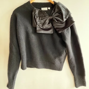 Svart stickad tröja med stor rosett - Supersnygg svart stickad tröja från Kappahl med en stor satinrosett framtill vid halsen. Tröjan har lång ärm och ribbade muddar vid ärmslut och nederkant. Använd 1 gång så är i ett bra skick💗