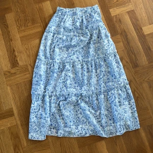 Blåvit blommig långkjol med volanger - Supersöt maxikjol i vitt med blått blommönster och flera volanglager. Kjolen har resår i midjan