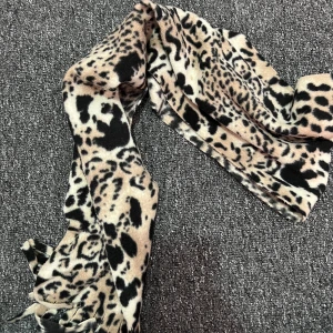 Leopardmönstrad beige halsduk  - Snygg halsduk med leopardmönster i beige, svart och bruna toner. Mjuk och varm med fransar i ändarna, perfekt att slänga över axlarna för en cool look. Passar dig som vill sticka ut lite extra i vinterkylan.