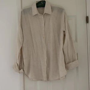 Beige linneskjorta med knappar från Nelly  - Stilren beige skjorta i linne med klassisk krage och knäppning framtill. Skjortan har långa ärmar som kan vikas upp för en avslappnad look. Perfekt för dig som gillar en enkel och tidlös stil.
