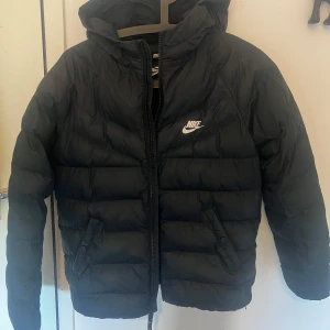 Svart pufferjacka från Nike barn - Svart pufferjacka från Nike med huva och dragkedja framtill. 