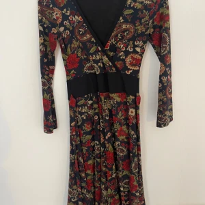 Blommig långklänning med v-ringning - Långklänning med v-ringning och lång ärm i mörkblått tyg med färgstarkt blommönster i rött, grönt och beige. Klänningen har omlottliv och markerad midja, vilket ger en snygg siluett. Perfekt för dig som gillar bohemiska vibes och mönster.
