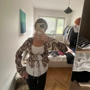 Brun mönstrad omlott overshirt - Säljer en snygg brun och beige overshirt med swirl-mönster och knytning framtill. Jackan har långa ärmar och croppad passform, perfekt för att lyfta vilken outfit som helst. Materialet känns mjukt och följsamt, och den unika designen gör den till ett riktigt statement-plagg.