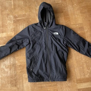 Svart vinter jacka The North Face DryVent - Svart fodrad vindjacka från The North Face med DryVent-teknologi. Jackan har huva, dragkedja framtill och två fickor med dragkedja. Insidan är fodrad med mjuk grå fleece för extra komfort. Perfekt för blåsiga dagar och outdoor vibes.