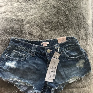 Lågmidjade jeansshorts från Gina Tricot - Snygga blå jeansshorts från Gina Tricot med låg midja och slitna detaljer. ALDRIG ANVÄNTS!!!Shortsen har fransig kant nertill, klassiska fickor fram och bak samt tydliga sömmar. Perfekta för en avslappnad och trendig look i sommar.